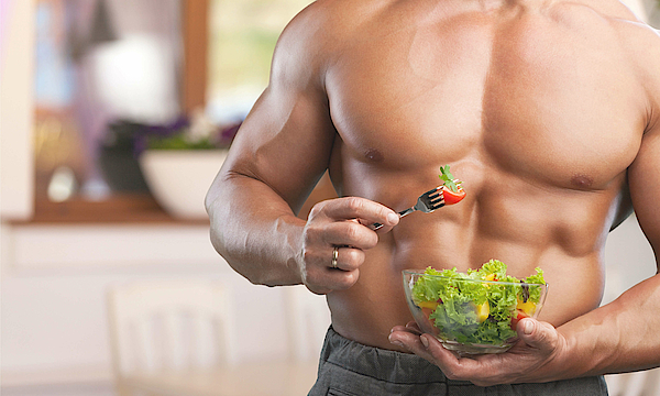 Ein Mann mit deutlich ausgeprägten Muskeln im Oberkörper hält eine Schüssel mit Salat. Er isst mit einer Gabel.