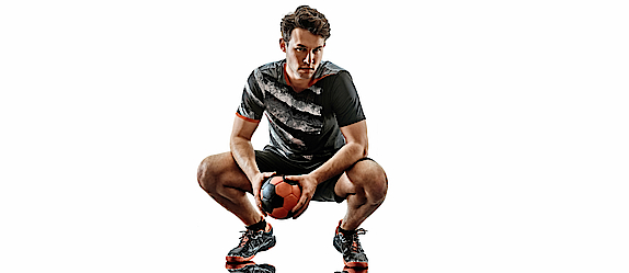 Ein Handballspieler in einem schwarzen Trikot ist in der Hocke. Er hält in seinen Händen einen Handball.