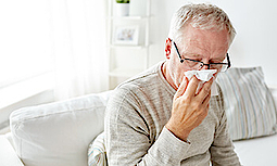 Ein älterer Herr mit Brille putzt sich die Nase. Er ist an Grippe erkrankt. Er sitzt auf einem Sofa.