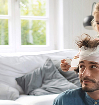 Das Mädchen verbindet mit einer weißen Mullbinde Vaters Kopf. Dabei sitzt er im Wohnzimmer auf dem Fußboden, das Mädchen steht hinter ihm. Auf dem Sofa sitzt ein kleiner Junge.