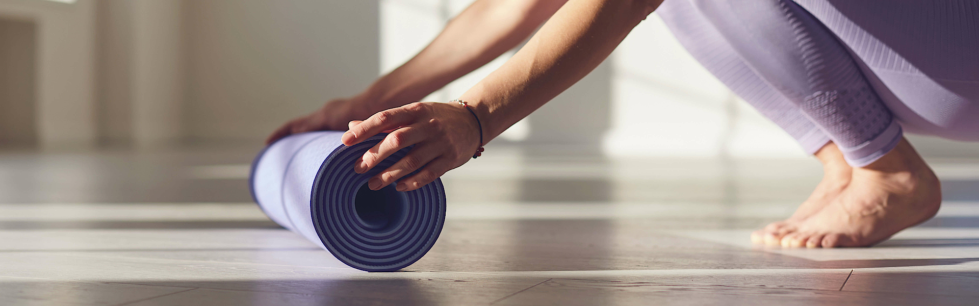 Eine Frau ist in der Hocke. Sie hat lilafarbene Sportkleidung an. Die Frau möchte eine Yogamatte ausrollen.