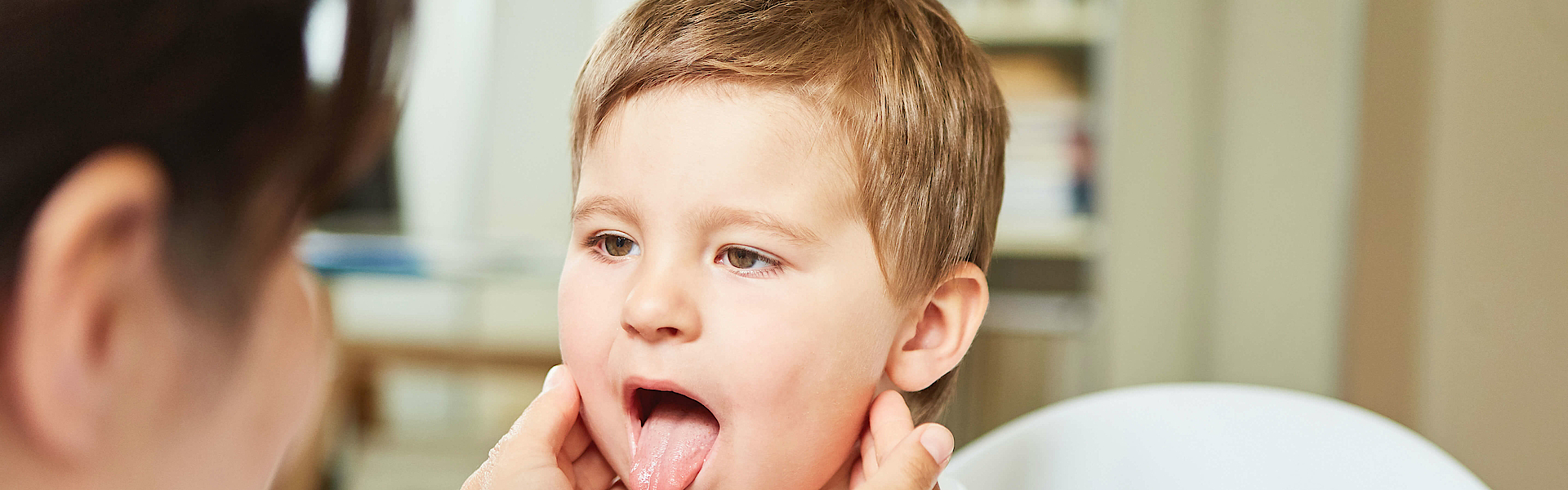 Ein Kinderarzt untersucht einen kleinen Jungen. Der Junge streckt die Zunge heraus. Der Arzt tastet den Hals ab.