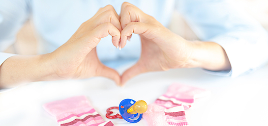 Mit den Fingern wird ein Herz symbolisiert, auf dem Tisch liegen Baby-Söckchen und ein Schnuller. 