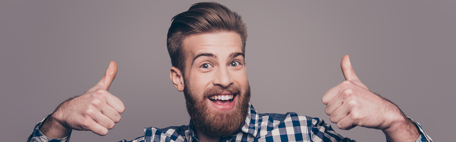 Mann mit Bart steht vor einer grauen Wand und hält beide Daumen hoch.
