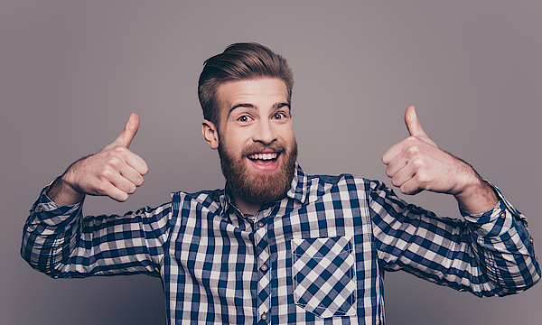 Ein Mann mit einem Bart und in karriertem Hemd steht vor einer grauen Wand. Er hält beide Daumen hoch zum ok und freut sich.