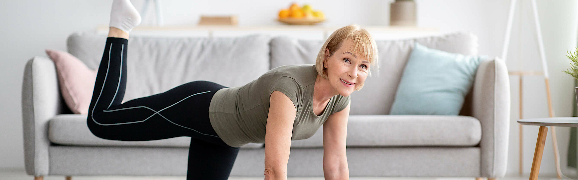 Frau macht im Wohnzimmer Gymnastik. Sie steht auf allen vieren und hebt ein Bein.