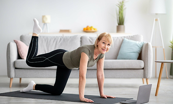 Frau macht im Wohnzimmer Gymnastik. Sie steht auf allen vieren und hebt ein Bein.