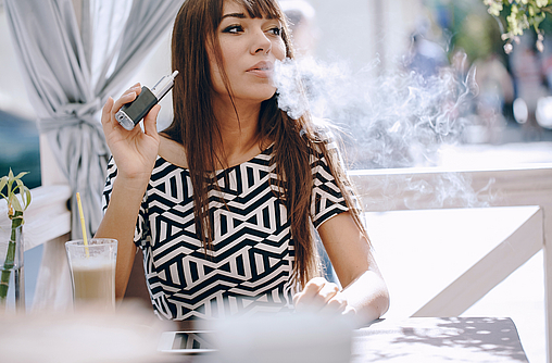 Schicke, dunkgelhaarige Frau sitzt an einem Tisch und ist beim Vapen.