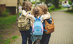 Drei kleine Schulkinder mit Rucksack laufen zur Schule. Das Mädchen läuft dabei in der Mitte und umarmt die Jungs.