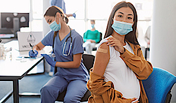 Eine Schwangere Frau hält ihren Arm für eine Impfung hin. Im Hintergrund sitzt eine Ärztin, welche einen Fragebogen ausfüllt. Beide tragen eine Schutzmaske