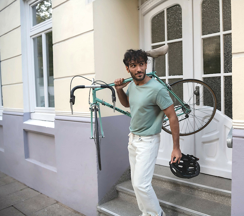 Ein Mann trägt rechts sein Fahrrad und steht auf einer Treppe. Er wendet den Blick nach links.