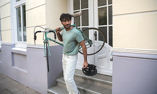 Ein Mann trägt rechts sein Fahrrad und steht auf einer Treppe. Er wendet den Blick nach links.