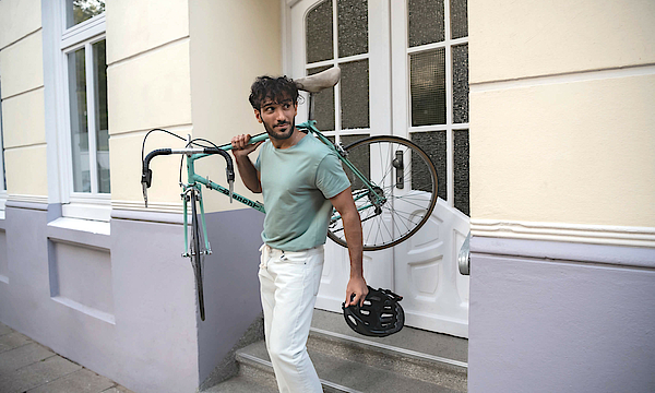 Ein Mann trägt rechts sein Fahrrad und steht auf einer Treppe. Er wendet den Blick nach links.