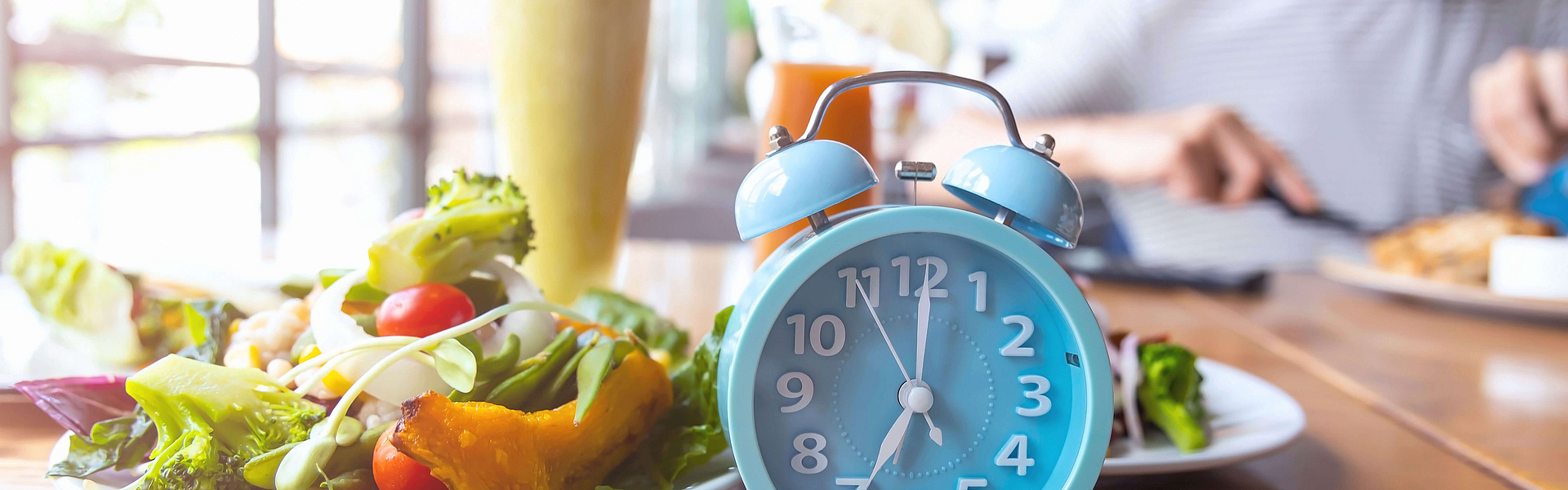 Auf einem Tisch steht ein blauer Wecker. Es wird kurz vor sieben Uhr angezeigt. Danebens teht ein Teller mit Gemüse. Im Hintergund ist ein Glas Orangensaft.
