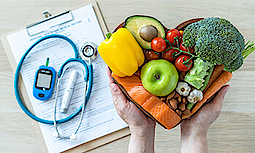 Ketogene Lebensmittel (Lachs, Avocado, Nüsse, Gemüse) in einer Holzschale in Herzform. Im Hintergrund liegen ein Fragebogen, Stethoskop und ein Blutzuckermessgerät.