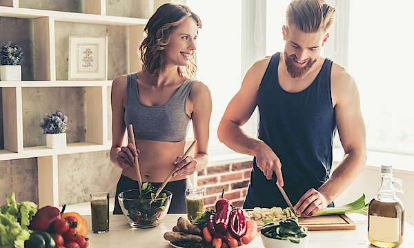 Ein Mann und eine Frau stehen in der Küche. Sie tragen Sportkleidung und kochen sich etwas.