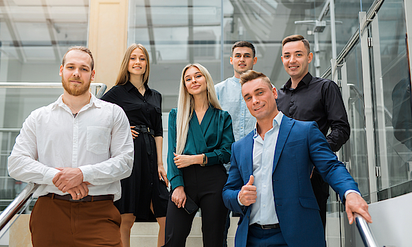 Eine Gruppe junger Menschen steht auf einer Treppe. Sie sind gut gekleidet. Sie haben ihren ersten Arbeitstag.