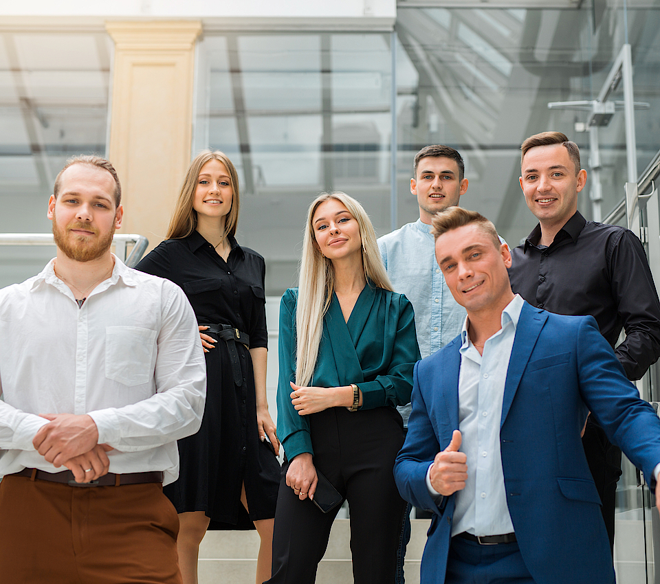 Eine Gruppe junger Menschen steht auf einer Treppe. Sie sind gut gekleidet. Sie haben ihren ersten Arbeitstag.