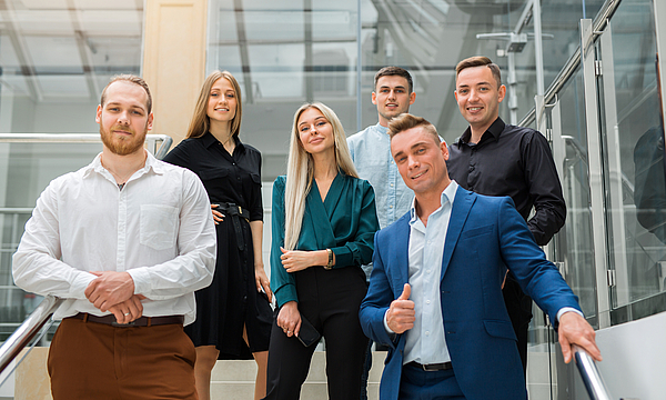 Eine Gruppe junger Menschen steht auf einer Treppe. Sie sind gut gekleidet. Sie haben ihren ersten Arbeitstag.