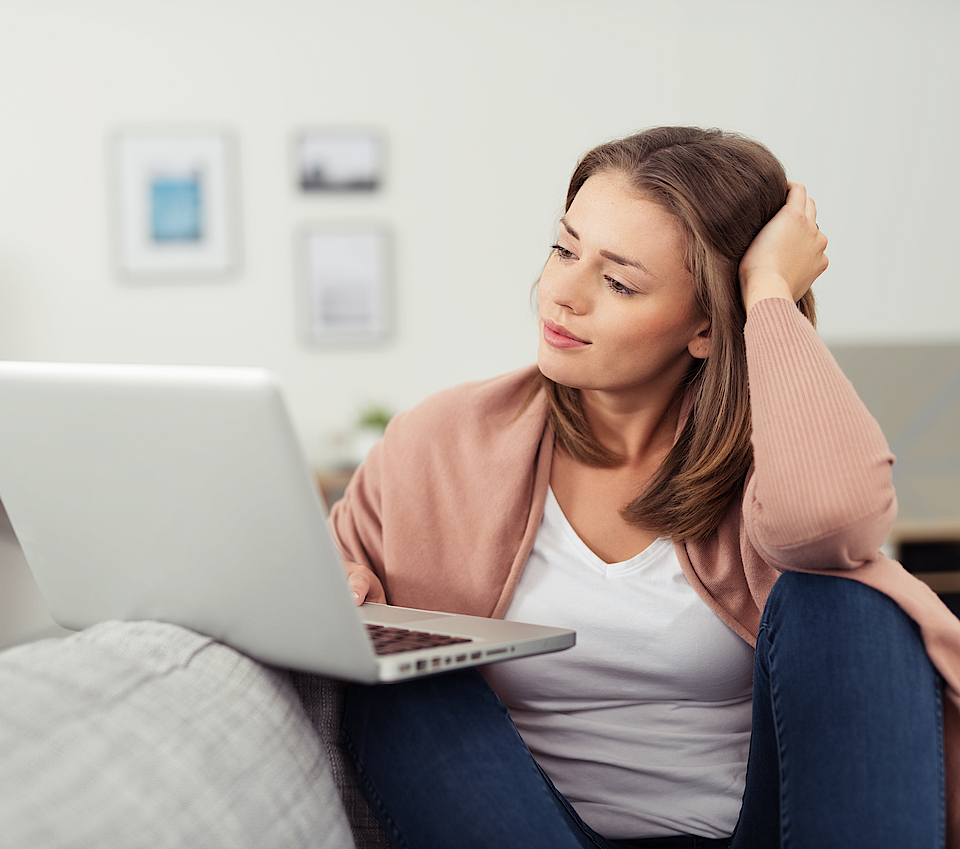Frau sitzt auf dem Sofa und arbeitet mit dem Laptop. Der Laptop ist auf dem Sofa abgelegt.