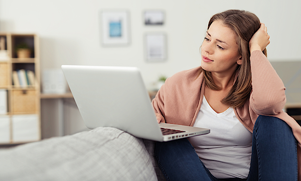 Frau sitzt auf dem Sofa und arbeitet mit dem Laptop. Der Laptop ist auf dem Sofa abgelegt.