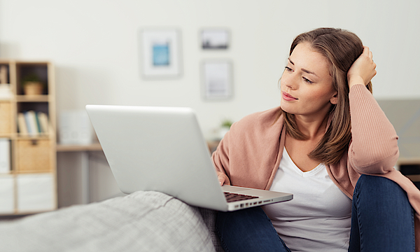 Frau sitzt auf dem Sofa und arbeitet mit dem Laptop. Der Laptop ist auf dem Sofa abgelegt.