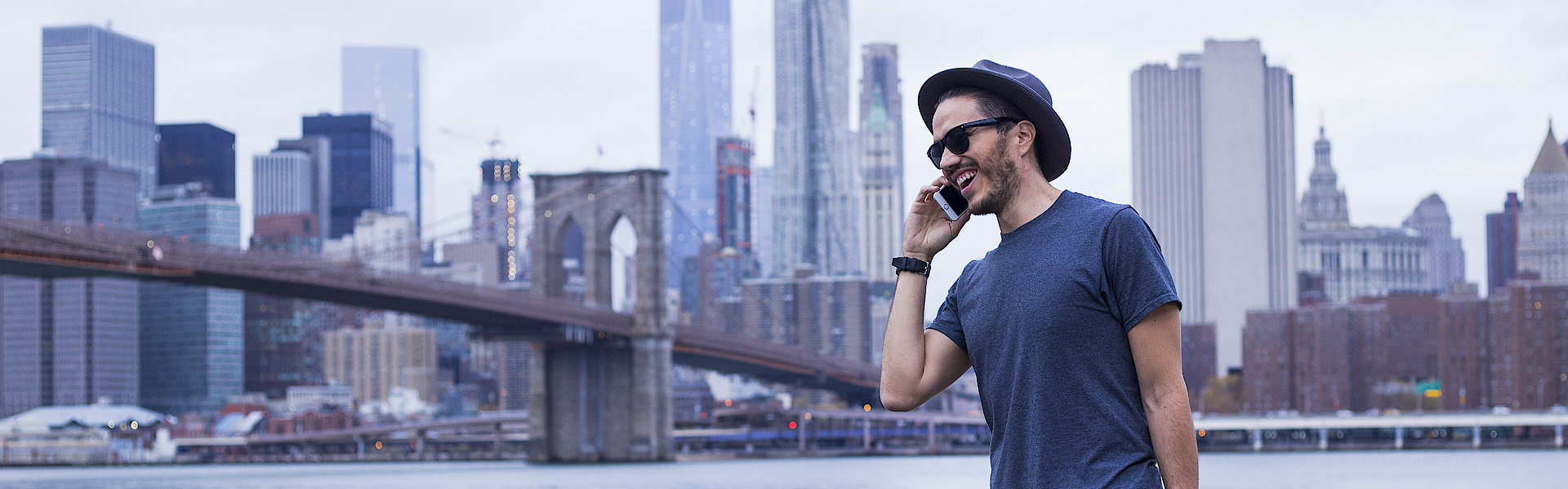 Ein Mann vor der Skyline von New York. Er trägt eine Laptop-Tasche und telefoniert.