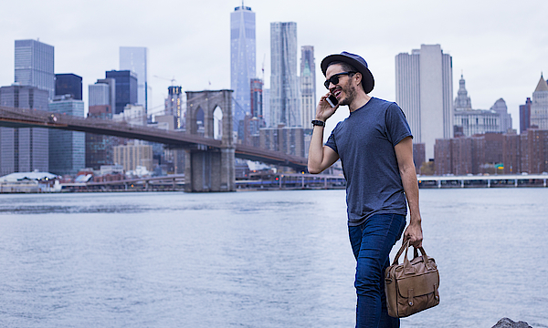 Ein Mann vor der Skyline von New York. Er trägt eine Laptop-Tasche und telefoniert.