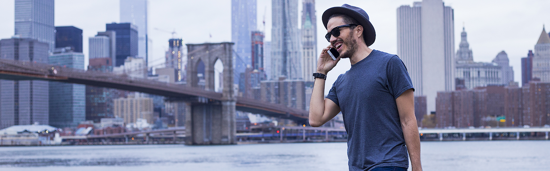 Ein Mann vor der Skyline von New York. Er trägt eine Laptop-Tasche und telefoniert.