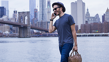 Ein Mann vor der Skyline von New York. Er trägt eine Laptop-Tasche und telefoniert.