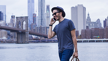 Ein Mann vor der Skyline von New York. Er trägt eine Laptop-Tasche und telefoniert.