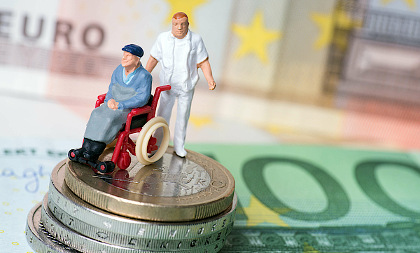 Ein Rollstuhlfahrer mit Pflegepersonal steht auf mehren Geldscheinen