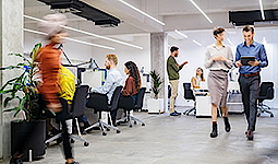 Menschen in einem modernen Büro. Manche Menschen sitzen an ihrem Schreibtisch, andere laufen. Zwei unterhalten sich dabei im Gehen. Zwei Menschen führen ein Gespräch im Sitzen und Stehen.