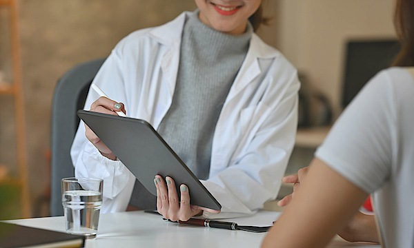 Ein Arzt und sein Patient sitzen an einem Tisch. Der Arzt hält ein Tablet in der Hand und zeigt darauf etwas dem Patienten.