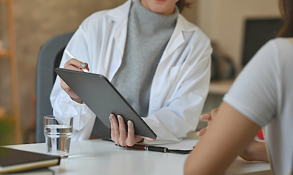 Ein Arzt und sein Patient sitzen an einem Tisch. Der Arzt hält ein Tablet in der Hand und zeigt darauf etwas dem Patienten.