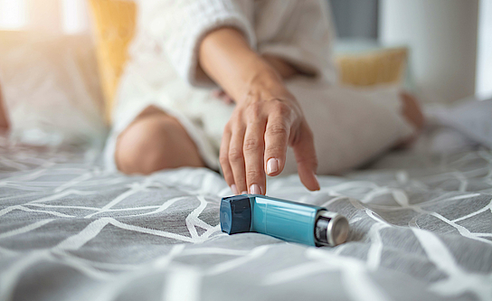 Auf einer grauen Decke liegt ein Inhalator. Eine weibliche Hand greift danach.