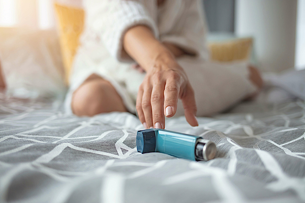 Auf einer grauen Decke liegt ein Inhalator. Eine weibliche Hand greift danach.