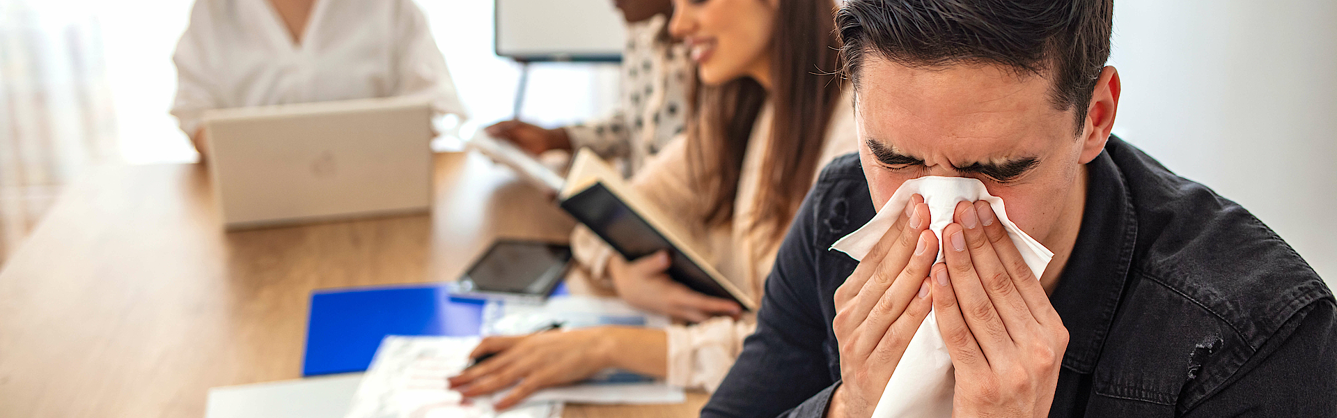 Der junge Mann bläst sich beim Arbeiten die Nase. Junge kranke Geschäftsleute, die sich während der Arbeit im Büro die Nase weht. Es sind Menschen im Hintergrund. Geschäftsmann, der sich die Nase mit Arbeitsgewebe zuschlägt