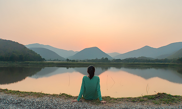 Eine Frau sitzt am Ufer eines Sees. Sie schaut aufs Wasser. Die Sonne geht unter. Sie ist alleine.