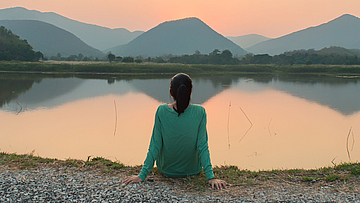 Eine Frau sitzt am Ufer eines Sees. Sie schaut aufs Wasser. Die Sonne geht unter. Sie ist alleine.