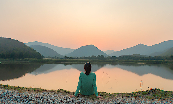 Eine Frau sitzt am Ufer eines Sees. Sie schaut aufs Wasser. Die Sonne geht unter. Sie ist alleine.
