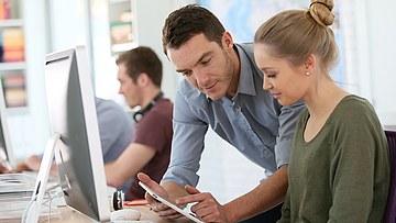 Ein Kollege erklärt einer junger Mitarbeiterin etwas am Computer.