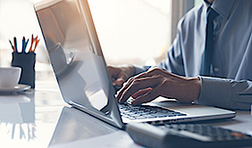 Nahaufnahme eines Mannes, der am Laptop arbeitet. Daneben liegt ein Taschenrechner