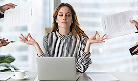 Eine Frau sitzt vor ihrem Laptop und meditiert. Es zeigen vier Hände in das Bild, welche Dokumente, ein Handy und einen Stift festhalten.