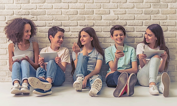 Jungen und Mädchen im Teenageralter verwenden technische Geräte, sprechen und lächeln, sitzen auf weißer Ziegelwand.