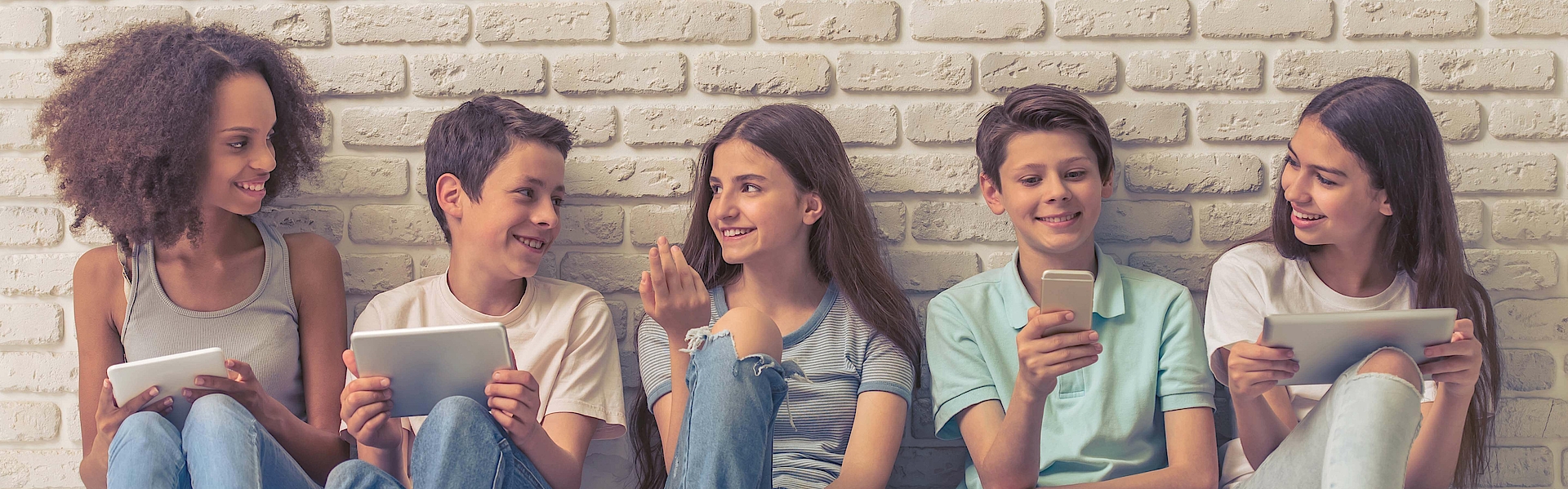 Jungen und Mädchen im Teenageralter verwenden technische Geräte, sprechen und lächeln, sitzen auf weißer Ziegelwand.