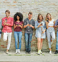 Eine Gruppe junger Menschen steht vor einer Mauer. Jeder hat ein Smartphone in der Hand und blickt darauf.