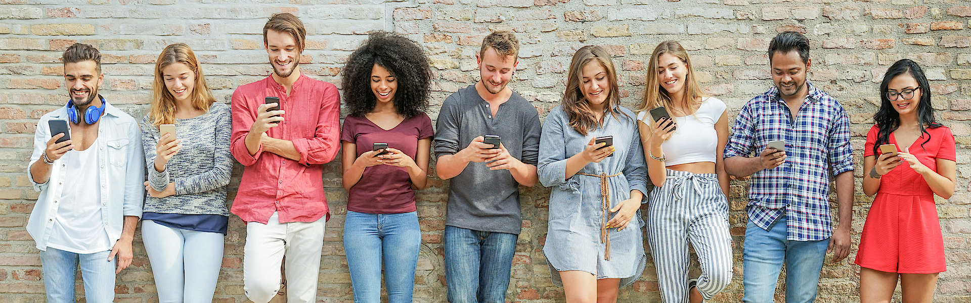 Eine Gruppe junger Menschen steht vor einer Mauer. Jeder hat ein Smartphone in der Hand und blickt darauf.