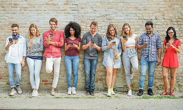 Eine Gruppe junger Menschen steht vor einer Mauer. Jeder hat ein Smartphone in der Hand und blickt darauf.