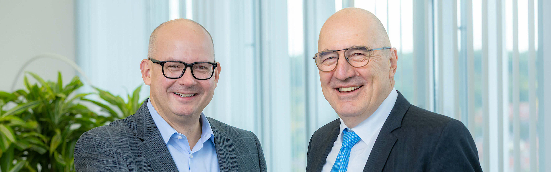 Links seht der neue Vorstand der mhplus, Heiko Kastner. Rechts steht der alte Vorstand der mhplus, Winfried Baumgärtner. Beide reichen sich die Hand.